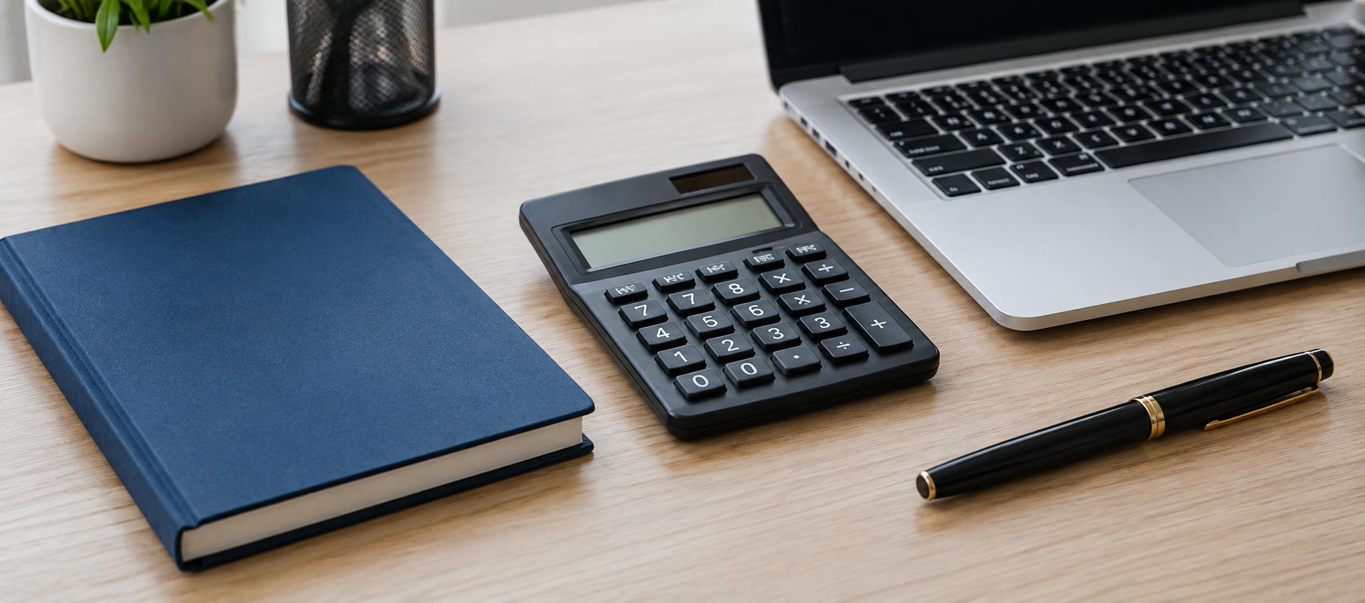Calculator, navy notebook, pen and laptop on an oak desk